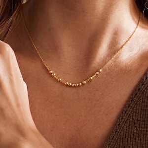 May include: A gold-toned necklace featuring a delicate chain with a central segment composed of alternating small round beads and short cylindrical bars, creating a linear pattern that rests along the collarbone in a subtle, curved arrangement.