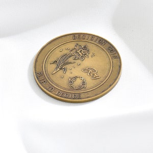 Puede incluir: Una moneda de color dorado con las palabras "Decision Coin" y "Flip to Decide" alrededor del borde. La moneda presenta una flor estilizada con un globo terráqueo y la palabra "Ray" en el centro.