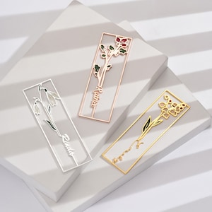 Może przedstawiać: Trzy złote metalowe zakładki do książek z różnymi wzorami kwiatowymi. Zakładki są obwiedzione złotem i mają białą, zieloną i czerwoną emaliowaną powierzchnię. Zakładki przedstawiają słowa "Ruelle", "Ruddie" i "Summer" napisane kursywą.