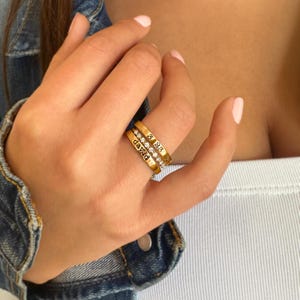 May include: Three gold rings are stacked on a finger. One ring is engraved with the name "david", another with the date "3 23 2018", and the third is encrusted with small, clear gemstones. The rings are displayed against a soft background.