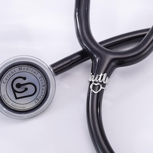 May include: A black stethoscope with a silver charm that says "faith" with a heart cutout. The stethoscope has a silver circular head with the text "Carent Medical Technology" and "Medical Stethoscope" on it.