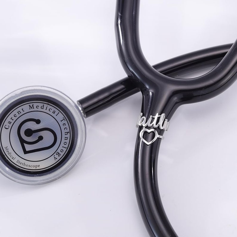 May include: A black stethoscope with a silver charm that says "faith" with a heart cutout. The stethoscope has a silver circular head with the text "Carent Medical Technology" and "Medical Stethoscope" on it.