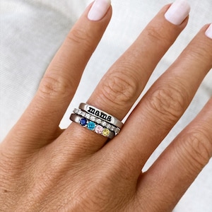 Pode incluir: Um conjunto de três anéis de prata empilhados num dedo. O anel superior tem a palavra "mama" gravada. O anel do meio é incrustado com pequenas pedras transparentes. O anel inferior tem quatro pedras preciosas coloridas: azul, azul claro, rosa e amarelo.