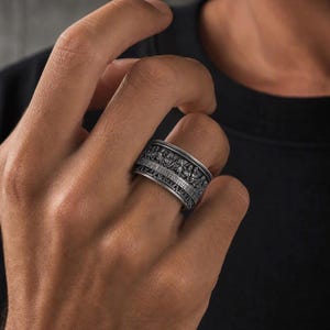 May include: A wide silver-toned metal ring featuring a detailed, embossed relief sculpture wrapping around the entire band. The design depicts a group of figures seated at a long table, rendered in a dark, oxidized finish to highlight the intricate textures and depth of the sculpted scene.