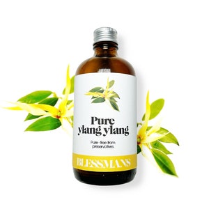 以下が含まれることがあります： 銀色の蓋が付いた茶色のガラス瓶で、「Pure ylang ylang」と「Pure-free from preservatives」のラベルが貼られ、その下にブランド名「Blessmans」が印刷されています。瓶は緑の葉のついた黄色の花に囲まれています。