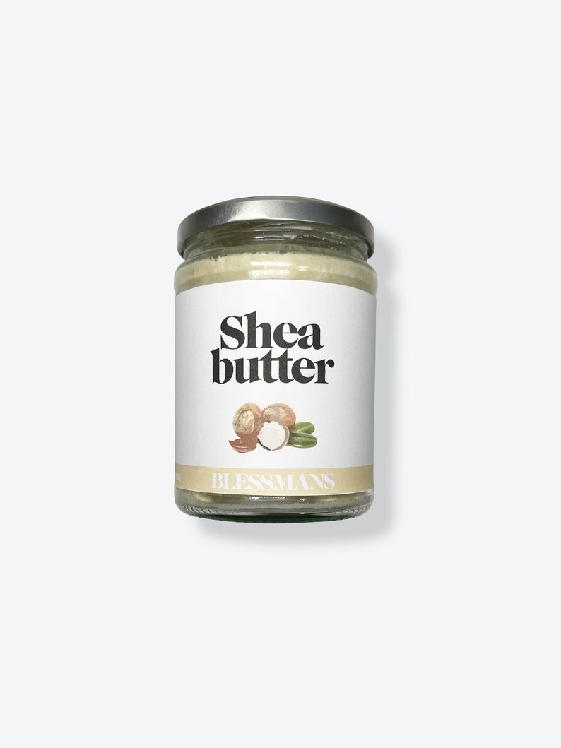 May include: A glass jar with a silver lid filled with shea butter. The label is white with black text that reads "Shea butter" and a drawing of shea nuts. The text "Blessmans" is printed in black at the bottom of the label.