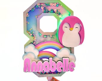 Puede incluir: Un topper de pastel de pingüino rosa con un arcoíris y nubes. El número 8 está decorado con lentejuelas iridiscentes y el nombre Annabelle está escrito en letras rosas.
