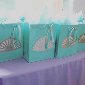 May include: Five teal gift bags with silver glitter seashell designs and pastel ribbon handles. The bags are arranged on a lavender tablecloth.