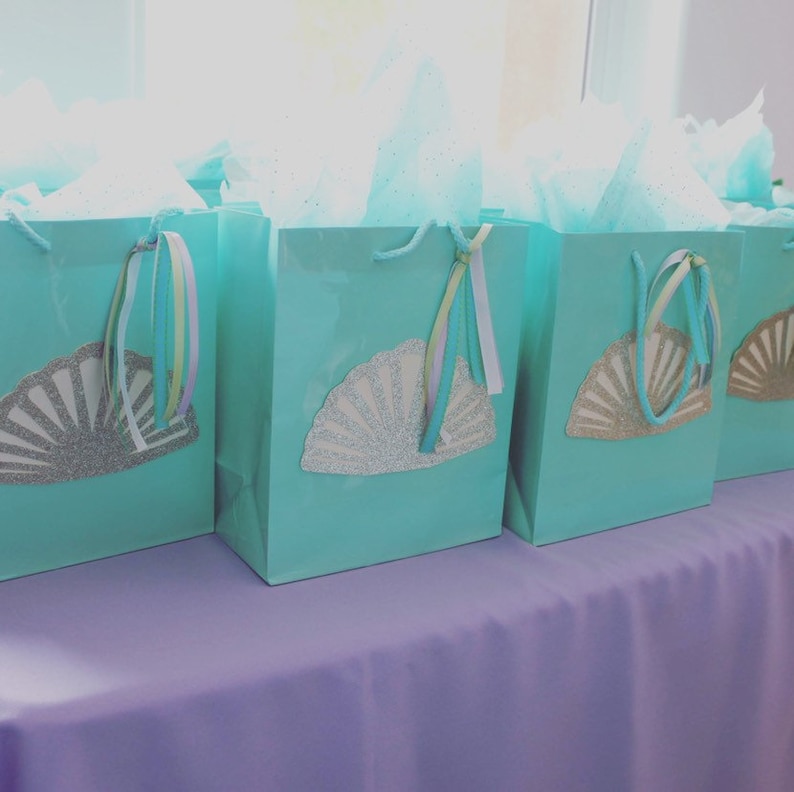 May include: Five teal gift bags with silver glitter seashell designs and pastel ribbon handles. The bags are arranged on a lavender tablecloth.