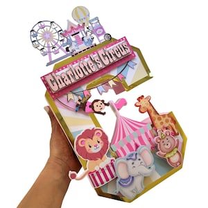 May include: A large, three-dimensional letter C decoration featuring a circus theme. It includes a Ferris wheel, a carousel, and a banner reading Charlotte's Circus. Adorned with cutouts of a monkey, giraffe, lion, elephant, and bear.