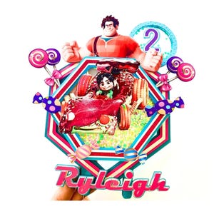 May include: A decorative cake topper featuring Wreck-It Ralph and Vanellope von Schweetz in a candy-themed race car. The design includes a blue circle with the number 2, colorful lollipops, wrapped candies, and the name Ryleigh in pink text.