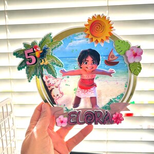 May include: A round cake topper featuring a cartoon character from the movie Moana. The character is a young girl with brown hair and brown eyes, wearing a red and white outfit. The topper also includes a pig, a rooster, a palm tree, a sun, and a boat. The name "Elora" is written on a pink banner at the bottom of the topper. The number "5" is also included on the topper.