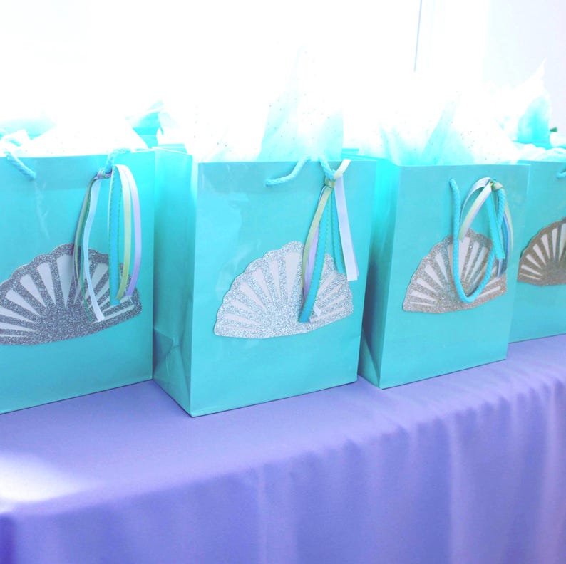 May include: Four turquoise blue gift bags with silver glitter seashell designs and pastel ribbon handles. The bags are arranged in a row on a lavender tablecloth.