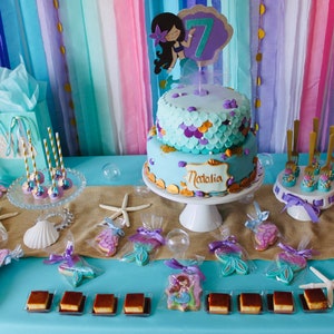May include: A mermaid-themed birthday party dessert table with a two-tiered cake decorated with purple, blue, and gold accents. The cake is topped with a mermaid figurine and the name "Natalia". The table is decorated with starfish, seashells, and mermaid-shaped cookies. There are also various other desserts, including cake pops, cupcakes, and chocolate squares.