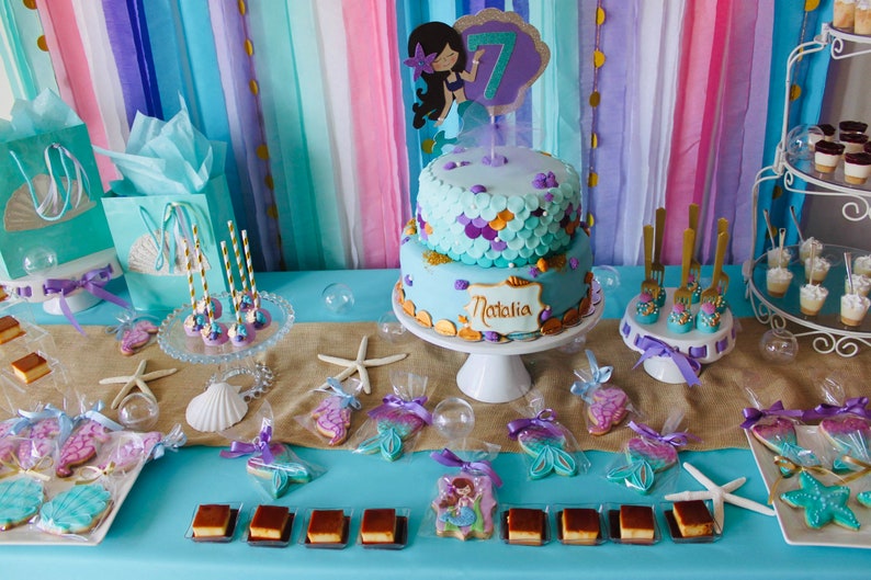 May include: A mermaid-themed birthday party dessert table with a two-tiered cake decorated with purple, blue, and gold accents. The cake is topped with a mermaid figurine and the name "Natalia". The table is decorated with starfish, seashells, and mermaid-shaped cookies. There are also various other desserts, including cake pops, cupcakes, and chocolate squares.