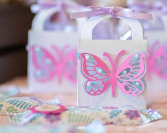 Puede incluir: Cajas de regalo blancas con diseño de mariposa y cinta morada. Las cajas están llenas de pequeños dulces y son perfectas para una fiesta o una ocasión especial.