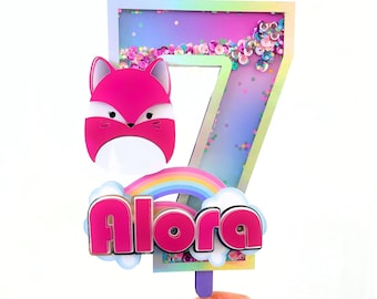 Puede incluir: Un adorno para pastel rosa y morado con un arcoíris, un zorro y el nombre "Alora" en letras rosas. El adorno está decorado con purpurina iridiscente.