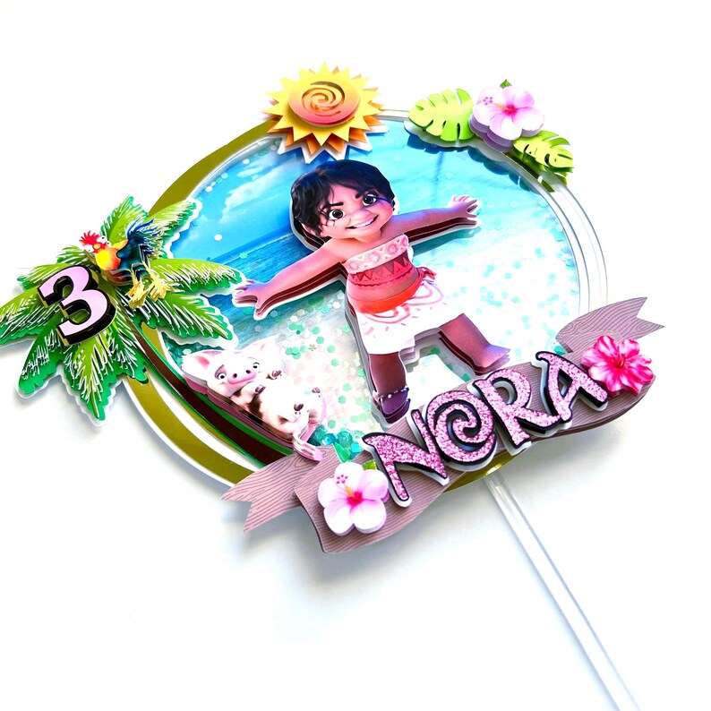 May include: A pink and green cake topper featuring a cartoon character from the movie Moana. The character is standing on a beach with a pig and a palm tree. The topper also includes the name "Nora" and the number "3".