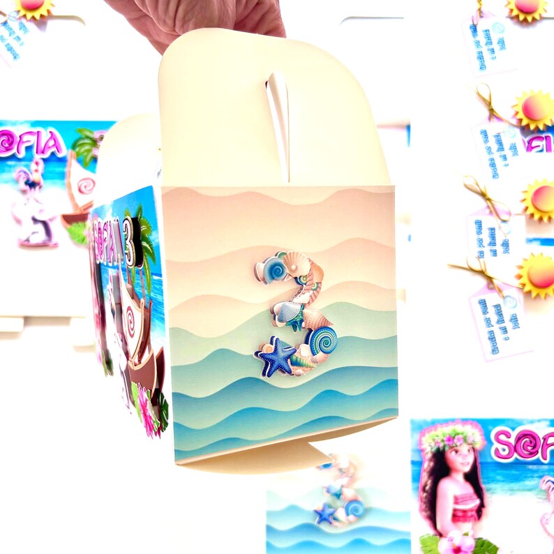 May include: A cream-colored party favor box with a handle, decorated with a beach-themed design. The box features a large number "3" made of seashells and a blue starfish. The background depicts ocean waves and a sandy beach.