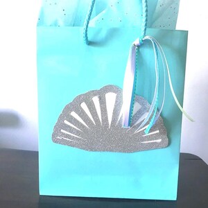 May include: A light blue gift bag with a silver glitter seashell design. The bag has a white and blue ribbon attached to the handle.
