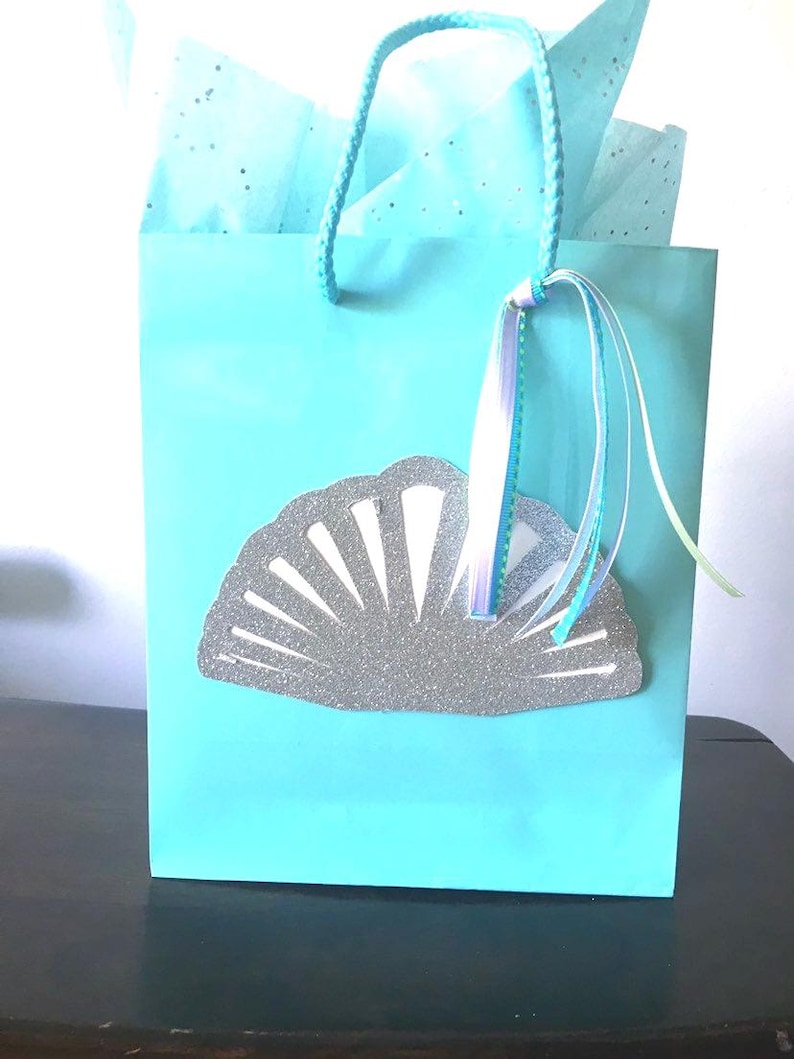 May include: A light blue gift bag with a silver glitter seashell design. The bag has a white and blue ribbon attached to the handle.