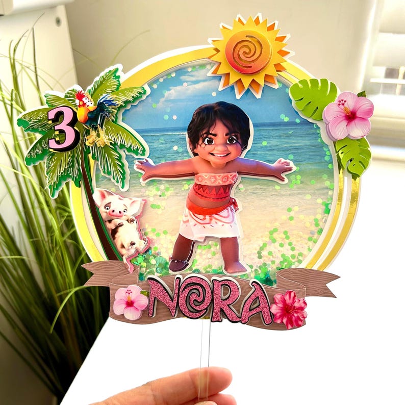 May include: A cake topper featuring a cartoon character from the movie Moana. The character is a young woman with brown hair and brown eyes, wearing a red and white dress. The topper also includes a palm tree, a pig, and the number 3. The name "Nora" is written on a banner at the bottom of the topper.