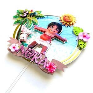 May include: A pink and gold cake topper featuring a cartoon character from the movie Moana. The character is standing on a beach with a pig and a palm tree. The topper says "Nora" and "3" on it.