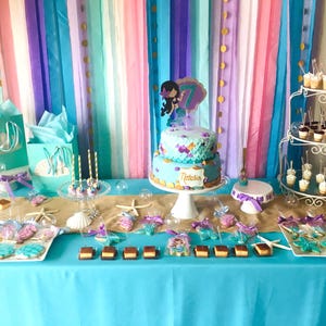 May include: A mermaid-themed birthday party dessert table with a two-tiered cake decorated with purple and blue frosting, a mermaid figurine, and the name "Natalia". The table is covered in a turquoise tablecloth and decorated with seashells, starfish, and other mermaid-themed decorations. There are also cupcakes, cookies, and other treats on the table.
