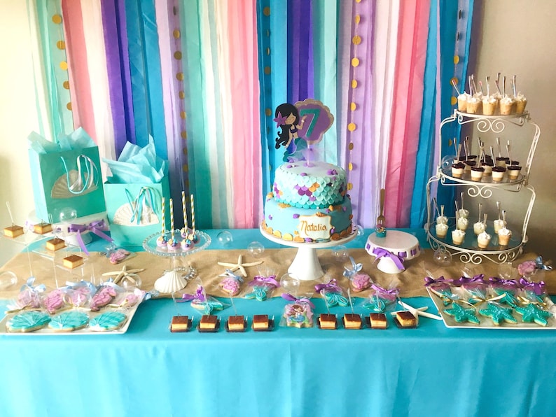 May include: A mermaid-themed birthday party dessert table with a two-tiered cake decorated with purple and blue frosting, a mermaid figurine, and the name "Natalia". The table is covered in a turquoise tablecloth and decorated with seashells, starfish, and other mermaid-themed decorations. There are also cupcakes, cookies, and other treats on the table.