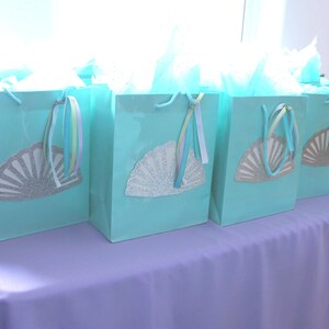 May include: A row of six turquoise gift bags with white tissue paper peeking out. Each bag has a white paper fan with silver glitter accents attached to the front.