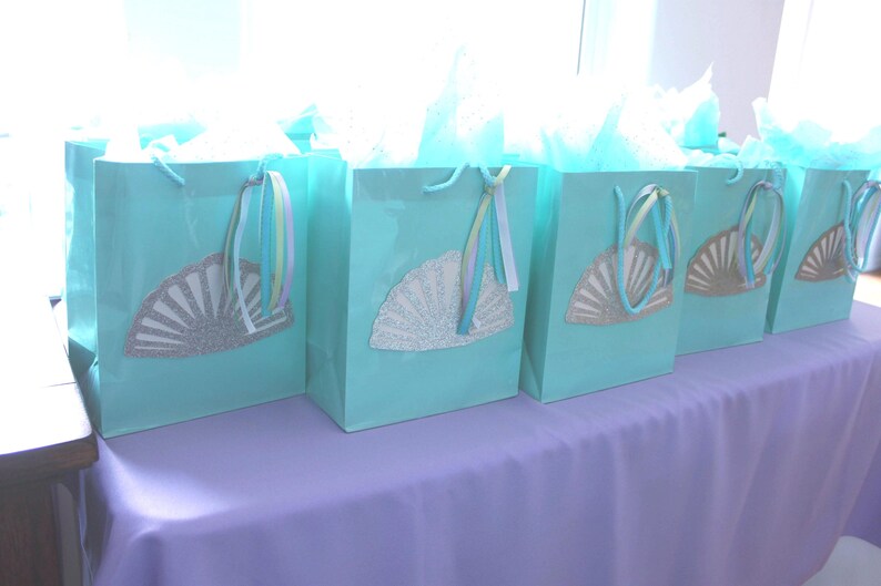 May include: A row of six turquoise gift bags with white tissue paper peeking out. Each bag has a white paper fan with silver glitter accents attached to the front.