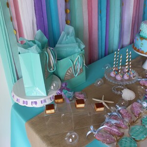 May include: A turquoise table with a burlap runner and a white cake stand with two teal gift bags. The table is decorated with teal and purple streamers, gold confetti, and a variety of desserts, including cupcakes, cookies, and cake pops.