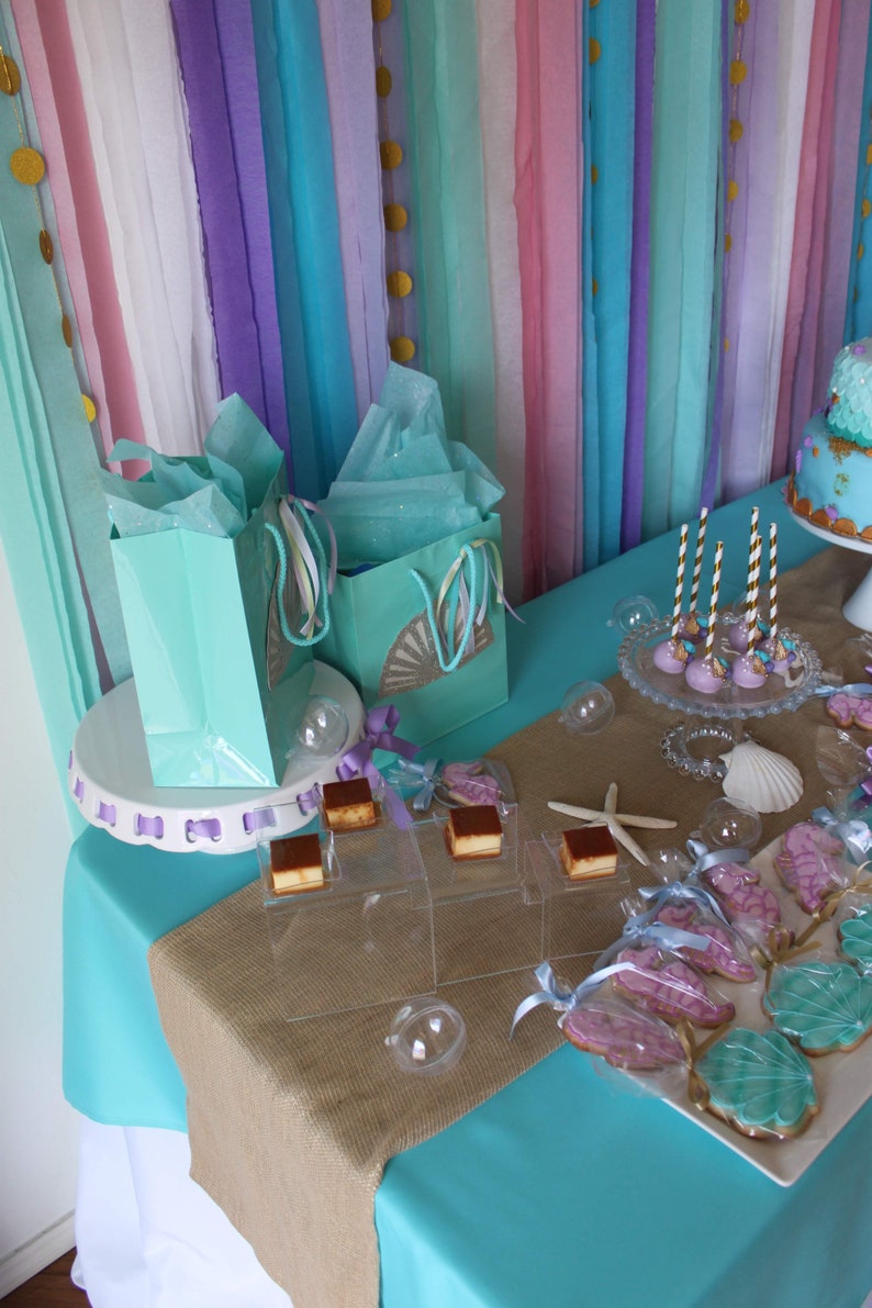 May include: A turquoise table with a burlap runner and a white cake stand with two teal gift bags. The table is decorated with teal and purple streamers, gold confetti, and a variety of desserts, including cupcakes, cookies, and cake pops.