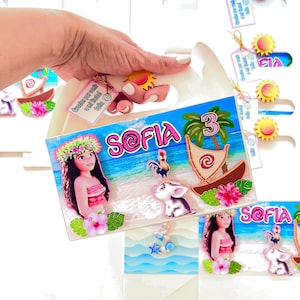 May include: A white gift box with a colorful Moana-themed design. The box features the name "Sofia" and the number "3", along with illustrations of Moana, a boat, and other tropical elements. Small tags with text are attached.