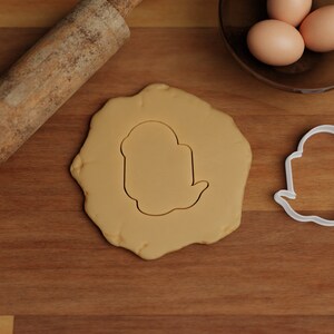 May include: A white cookie cutter shaped like a ghost on a wooden surface with a rolling pin and a bowl of eggs in the background.