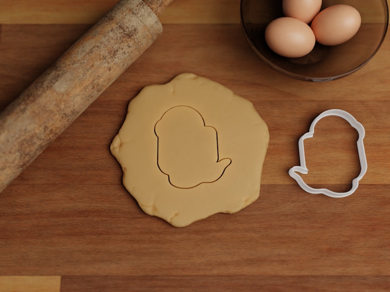 May include: A white cookie cutter shaped like a ghost on a wooden surface with a rolling pin and a bowl of eggs in the background.