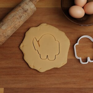 May include: A white plastic cookie cutter shaped like a character from the video game Among Us. The cookie cutter is on a wooden surface next to a rolling pin and a bowl of eggs.