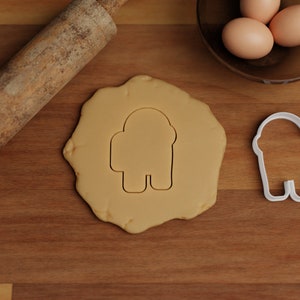 May include: A white plastic cookie cutter shaped like an astronaut, a rolling pin, and a ball of cookie dough on a wooden surface.