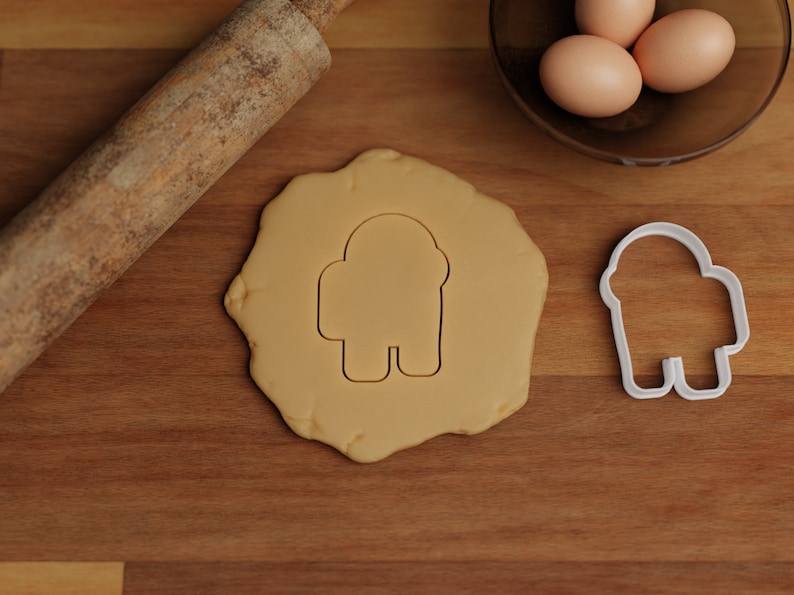May include: A white plastic cookie cutter shaped like an astronaut, a rolling pin, and a ball of cookie dough on a wooden surface.