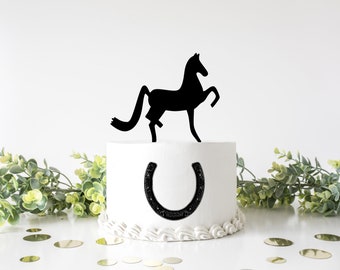 Könnte beinhalten: Ein weißer Kuchen mit schwarzem Frosting und einer schwarzen Silhouette eines Pferdes oben drauf. Der Kuchen ist mit einem schwarzen Hufeisen auf der Vorderseite verziert.