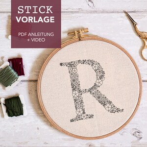 以下が含まれることがあります： 小さな花の模様でできた「R」の文字が刺繍された、完成した刺繍枠のクローズアップ。枠は、緑と赤紫の2つの刺繍糸の糸巻きと、金色の刺繍はさみと一緒に、白い木製の表面の上に置かれています。