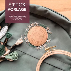 Puede incluir: Una mochila de lona verde con un parche de cuero marrón que dice "Beckmann Norway 1946". El parche está rodeado de flores bordadas. Hay varios carretes de hilo de bordar de varios colores en el lado izquierdo de la imagen.