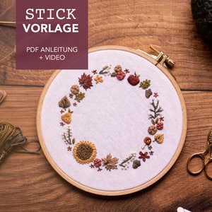 May include: A close-up of a white embroidery hoop with a brown wooden frame. The hoop is filled with a colorful embroidery design of a wreath with leaves, flowers, and berries. The text "STICK VORLAGE" is at the top of the image, and the text "PDF ANLEITUNG + VIDEO" is below the hoop.