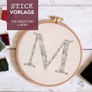 Könnte beinhalten: Nahaufnahme eines Reifens mit weißem Stoffhintergrund und einem schwarzen Stickmuster des Buchstabens "M" aus kleinen Blumen. Der Reifen liegt auf einer Holzoberfläche mit drei Spulen Stickgarn.