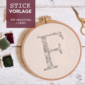 Könnte beinhalten: Ein hölzerner Stickrahmen mit einem weißen Stoff, der darüber gespannt ist. Der Stoff hat ein schwarzes Stickmuster des Buchstabens F, das aus kleinen Blumen besteht. Zwei Spulen Stickgarn, eine grüne und eine burgunderrote, liegen auf einer weißen Oberfläche neben dem Rahmen. Eine goldene Schere befindet sich rechts neben dem Rahmen. Der Text "STICK VORLAGE" befindet sich oben im Bild, und der Text "PDF ANLEITUNG + VIDEO" befindet sich unter dem Rahmen.