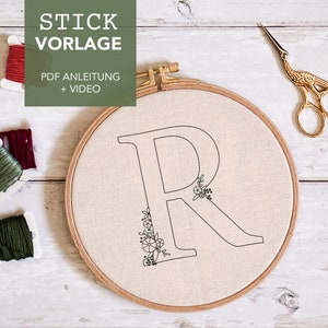Könnte beinhalten: Nahaufnahme eines weißen Stickrahmens mit einer schwarzen Umrandung des Buchstabens "R" mit floralen Details. Der Rahmen liegt auf einer weißen Holzoberfläche mit drei Spulen Stickgarn, einer roten, einer burgunderroten und einer grünen, sowie einer goldenen Stickerschere.