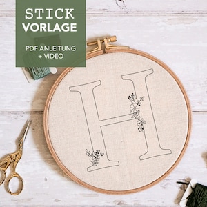 Könnte beinhalten: Ein Stickrahmen mit beigem Stoff, der einen schwarzen Umriss des Buchstabens "H" mit floralen Elementen zeigt. Der Text "STICK VORLAGE PDF ANLEITUNG + VIDEO" befindet sich in der oberen linken Ecke. Goldscheren und Garnrollen sind ebenfalls sichtbar.
