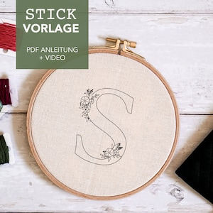 Könnte beinhalten: Nahaufnahme eines weißen Stickrahmens mit einer schwarzen Umrandung des Buchstabens "S" mit floralen Verzierungen. Der Rahmen befindet sich auf einer weißen Holzoberfläche mit drei Spulen Stickgarn.
