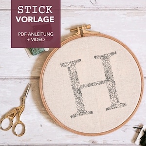 Könnte beinhalten: Nahaufnahme eines fertigen Stickrahmens mit dem Buchstaben "H" in schwarzem Garn gestickt. Der Buchstabe besteht aus kleinen floralen Designs. Der Rahmen befindet sich auf einer weißen Holzoberfläche mit Stickgarn und einer Schere.