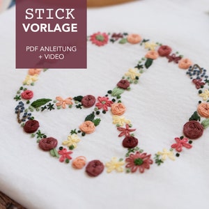 May include: A white fabric with a floral embroidery design of a peace sign. The embroidery is done in shades of pink, red, and yellow. The text "STICK VORLAGE" is in a maroon rectangle at the top of the image. The text "PDF ANLEITUNG + VIDEO" is below the first text.